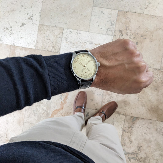 Deutsche Präzisions-Uhrenfabrik Glashütte Armbanduhr mit cremefarbenem Zifferblatt und schwarzem Armband, auf einem Handgelenk über Fliesenboden gezeigt.