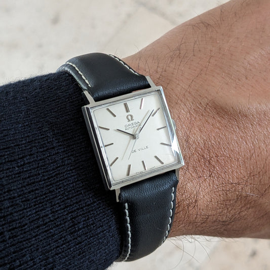 Omega De Ville Automatik 161.014 am Handgelenk, zeigt quadratisches silberfarbenes Gehäuse, helles Zifferblatt, schwarzes genähtes Armband und Außenhintergrund.