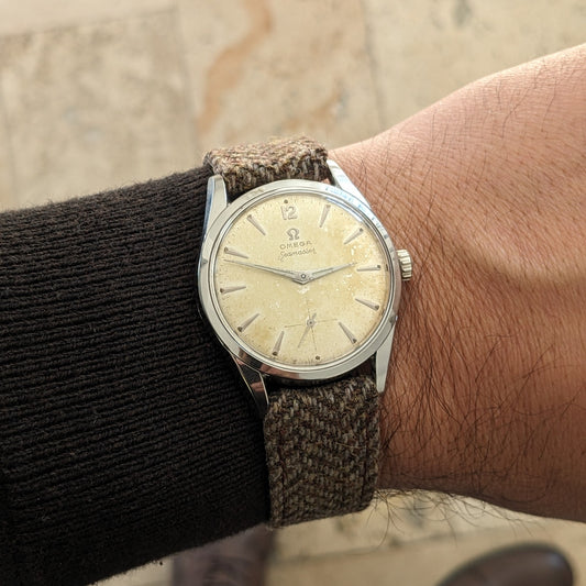 Omega Seamaster रेफरेंस 29374 कलाई पर पहना हुआ, क्रीम डायल, पतला पॉलिश्ड केस, बुना हुआ स्ट्रैप और काली आस्तीन दिखा रहा है।