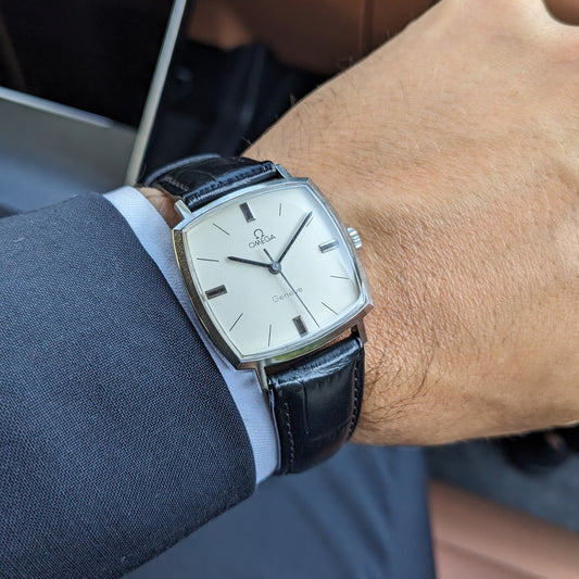Omega Genève reference 131.022 worn on a wrist, showing the cream square dial, silver case, and black leather strap over a blue shirt cuff.