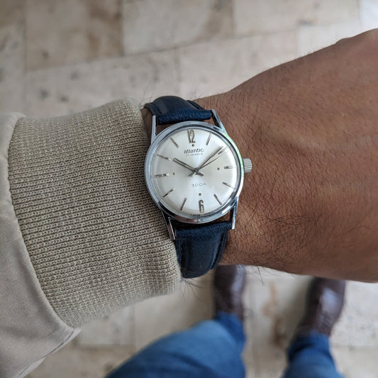 Atlantic 3004 portée au poignet avec une manche beige, bracelet en cuir bleu et cadran argenté, photographiée sur un sol à motif pierre.