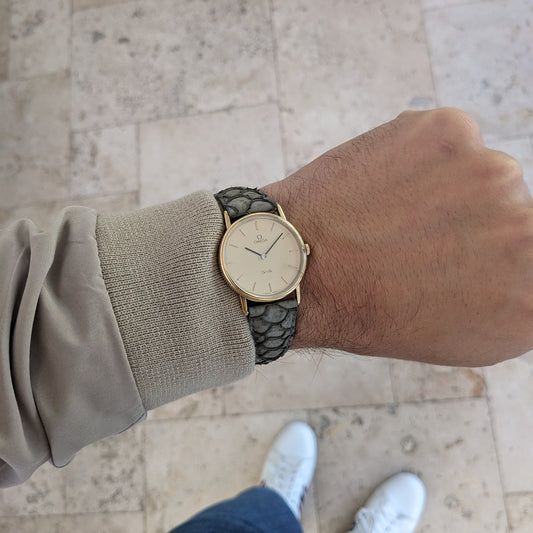 Wrist view of the Omega De Ville reference 111 0156 with cream dial and gray strap, photographed above a pale stone walkway.