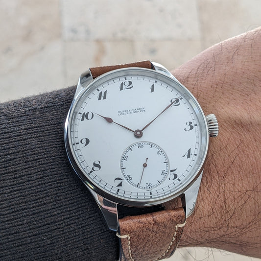 Ulysse Nardin कलाई घड़ी कलाई पर पहनी हुई, इसकी चमकदार केस, सफेद अंक डायल, छोटे सेकंड रजिस्टर और भूरे पट्टे के साथ।