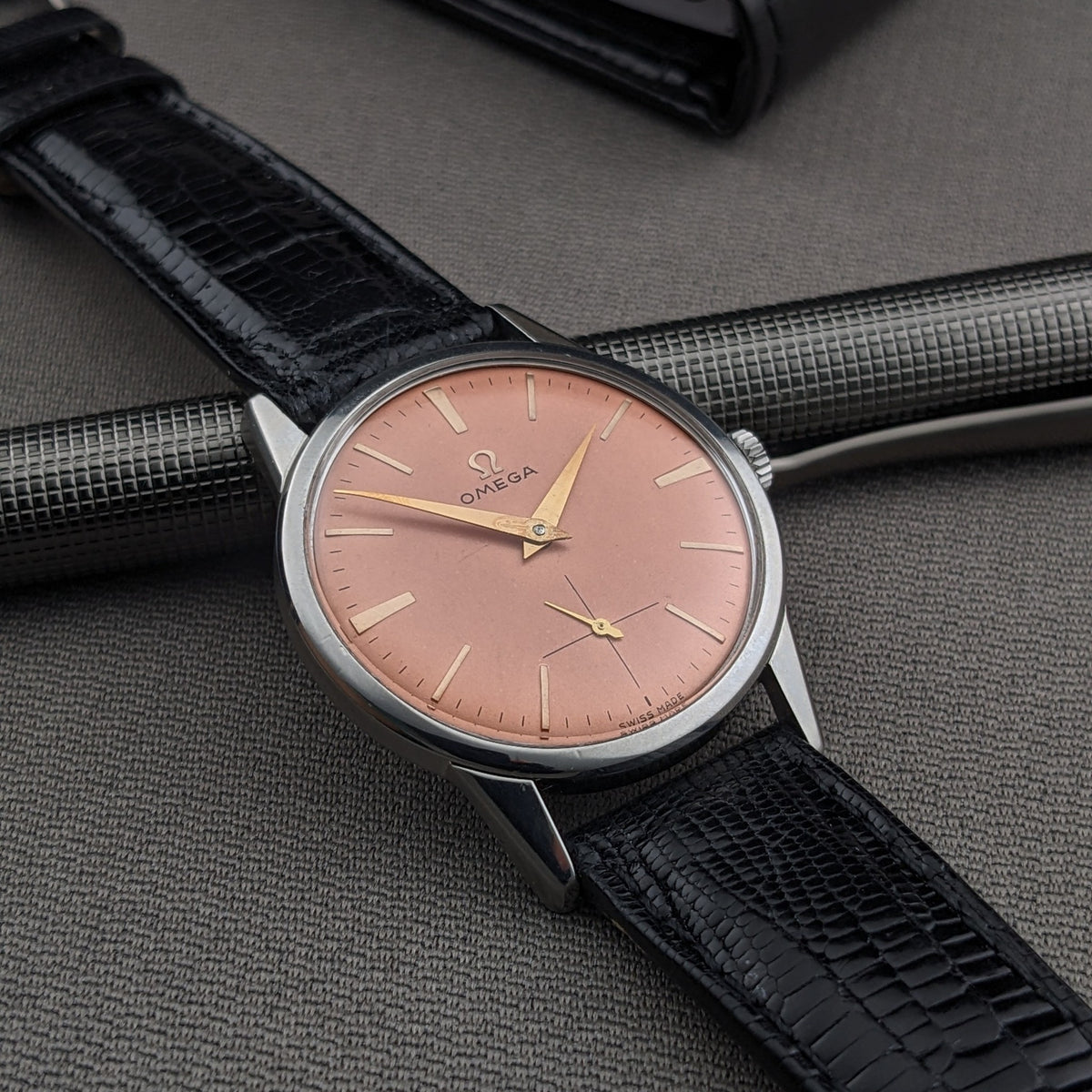 Orologio da polso Omega Classic referenza 14391-1 in diagonale con quadrante rosa, cinturino nero, cassa argento e penna su tessuto grigio.