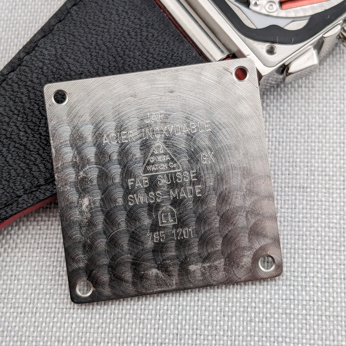 Close-up of an engraved metal plate beside the Omega Constellation Quadra 1841.73.31, with a black and red strap visible behind it.