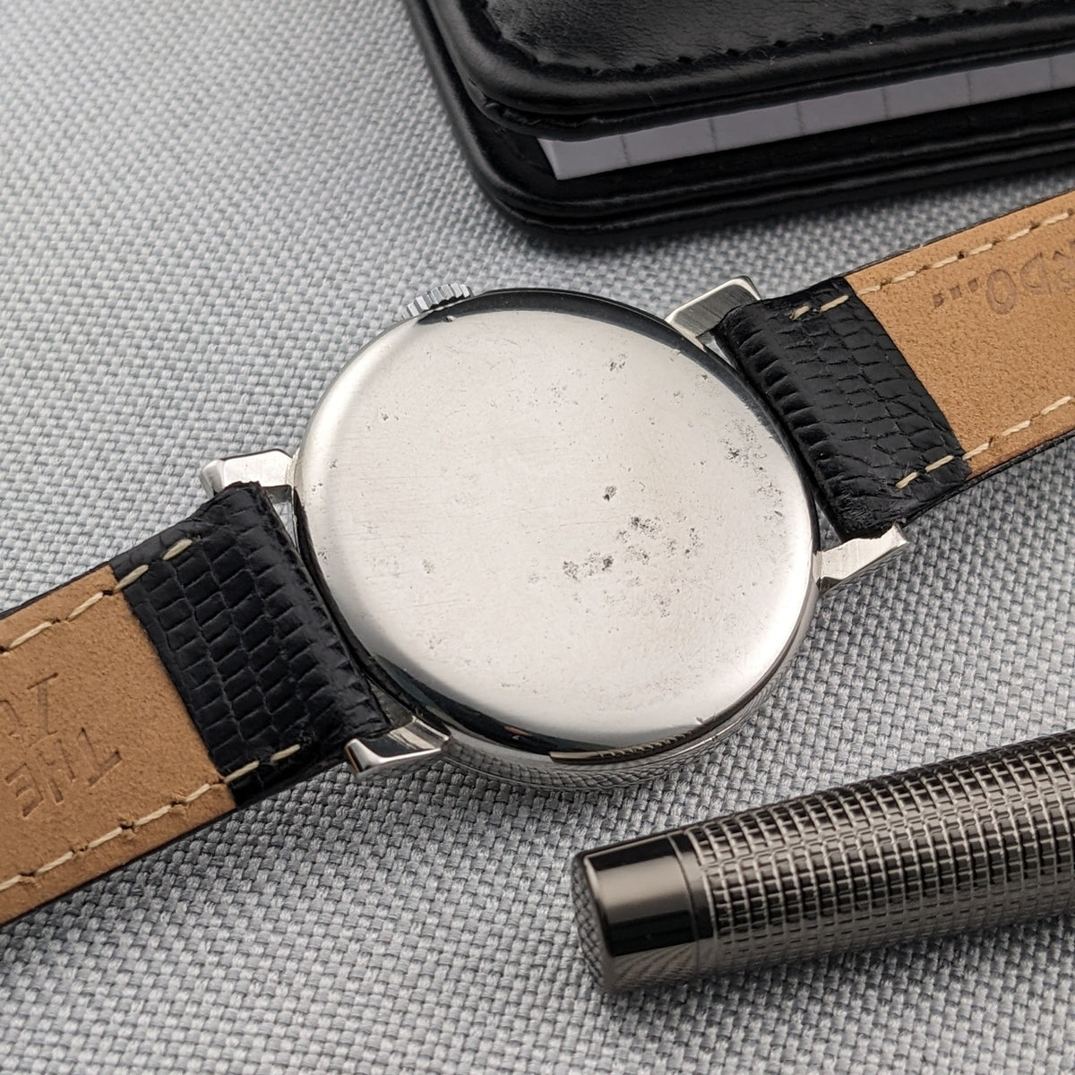Omega watch shown from the back with a plain round case, black textured strap, black wallet and metal pen on gray fabric.
