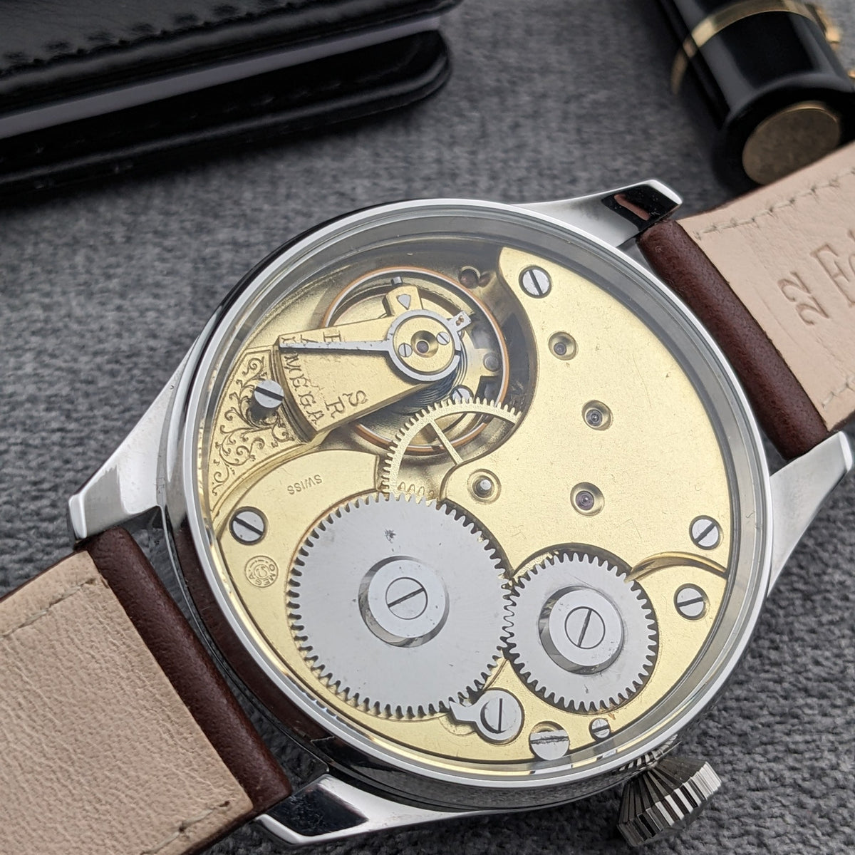 Orologio Omega visto dal retro trasparente, con movimento meccanico dorato, ingranaggi esposti, viti, cinturino marrone e corona visibili.