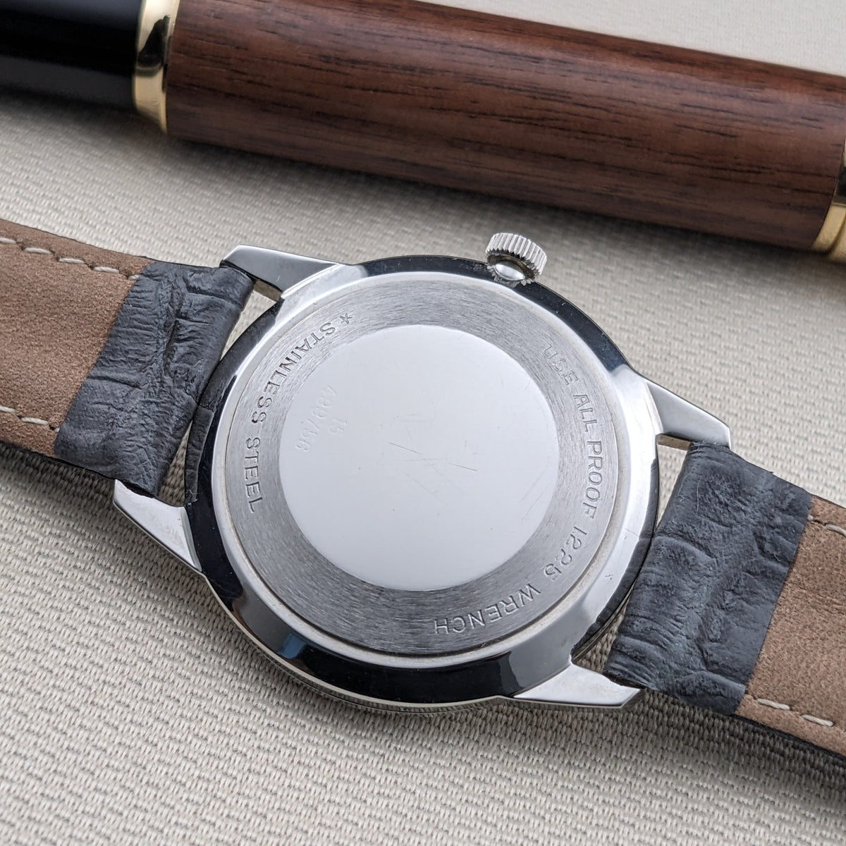 Longines Ultra-Chron 7827 shown from the rear on a black leather strap, with engraved caseback text and a wooden pen above.