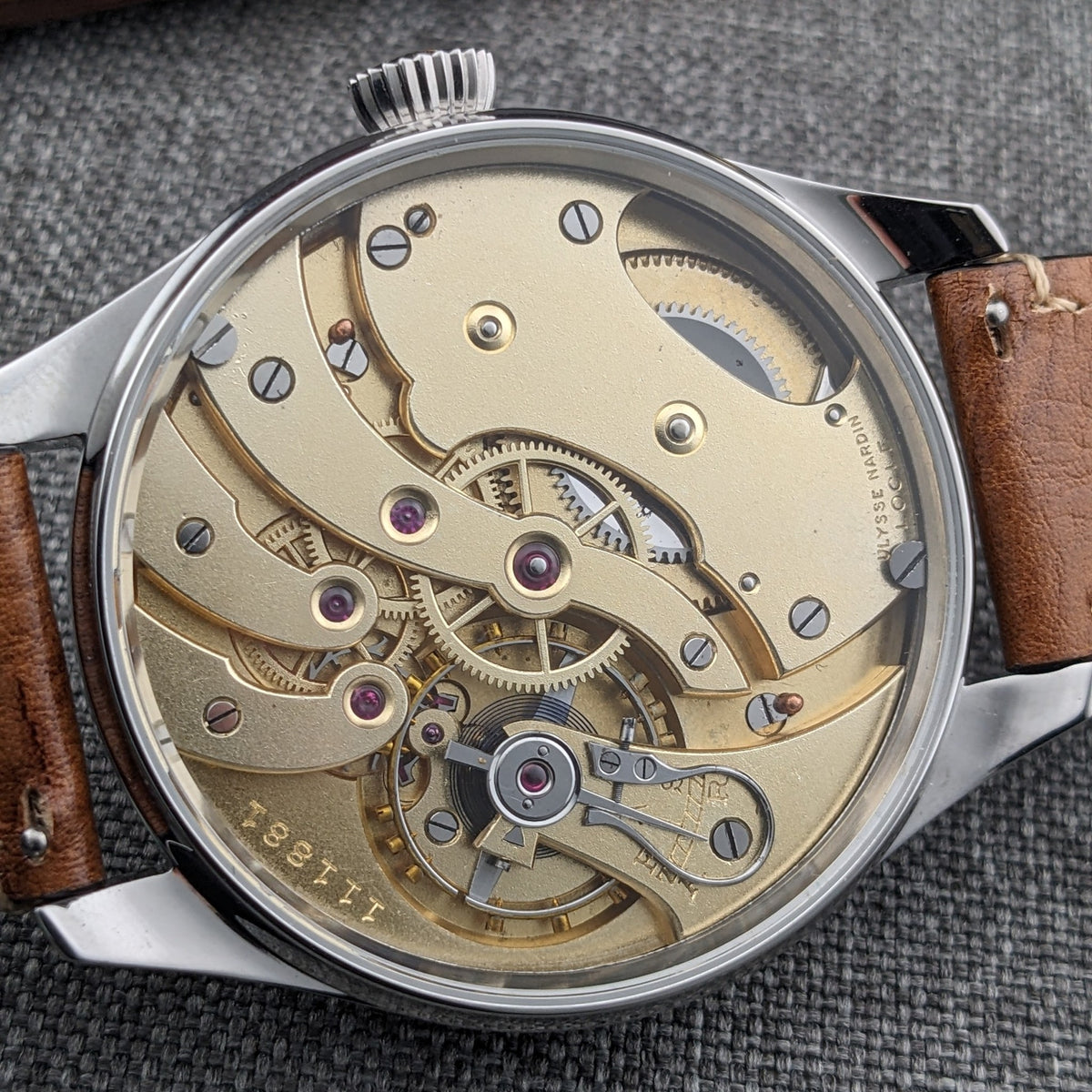 Ulysse Nardin घड़ी पीछे से दिखाई गई, जिसमें सुनहरे रंग का यांत्रिक मूवमेंट, दृश्य गियर्स, रूबी रंग के रत्न और उत्कीर्ण घटक हैं।