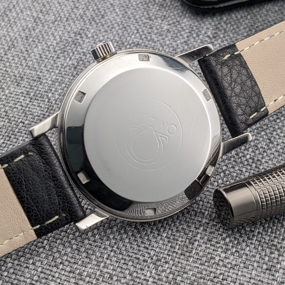 Omega Geneve Automatic reference 166.0163 shown from the rear, with engraved Omega emblem, polished caseback, black leather strap and gray woven backdrop.