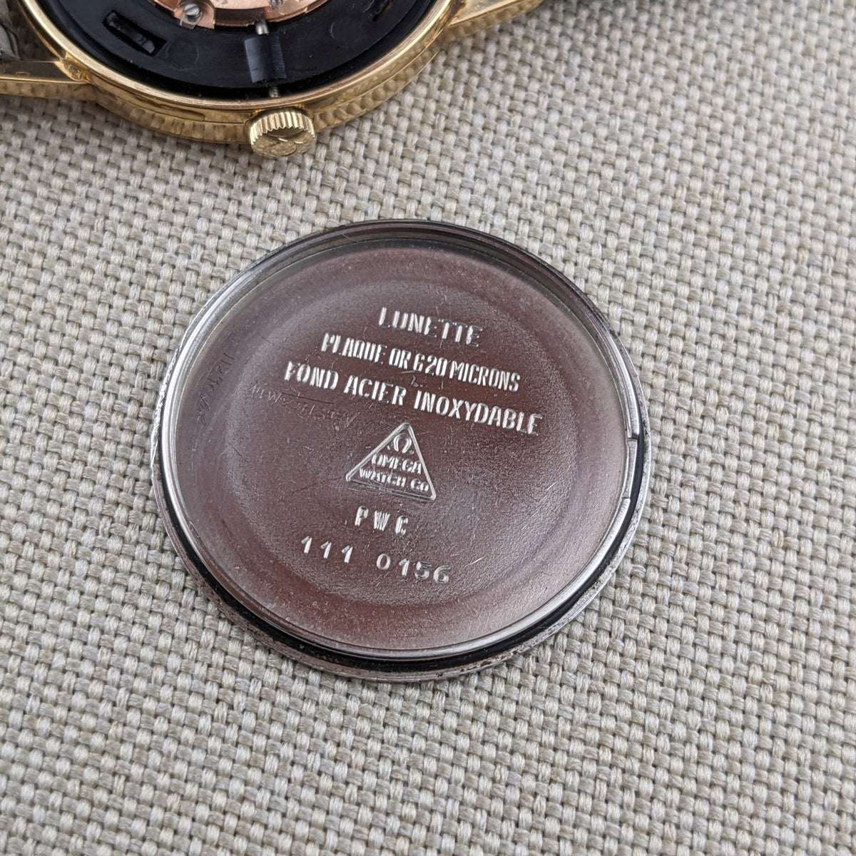Omega De Ville reference 111 0156 caseback with engraved text and triangular emblem, beside the gold-tone watch case on woven fabric.