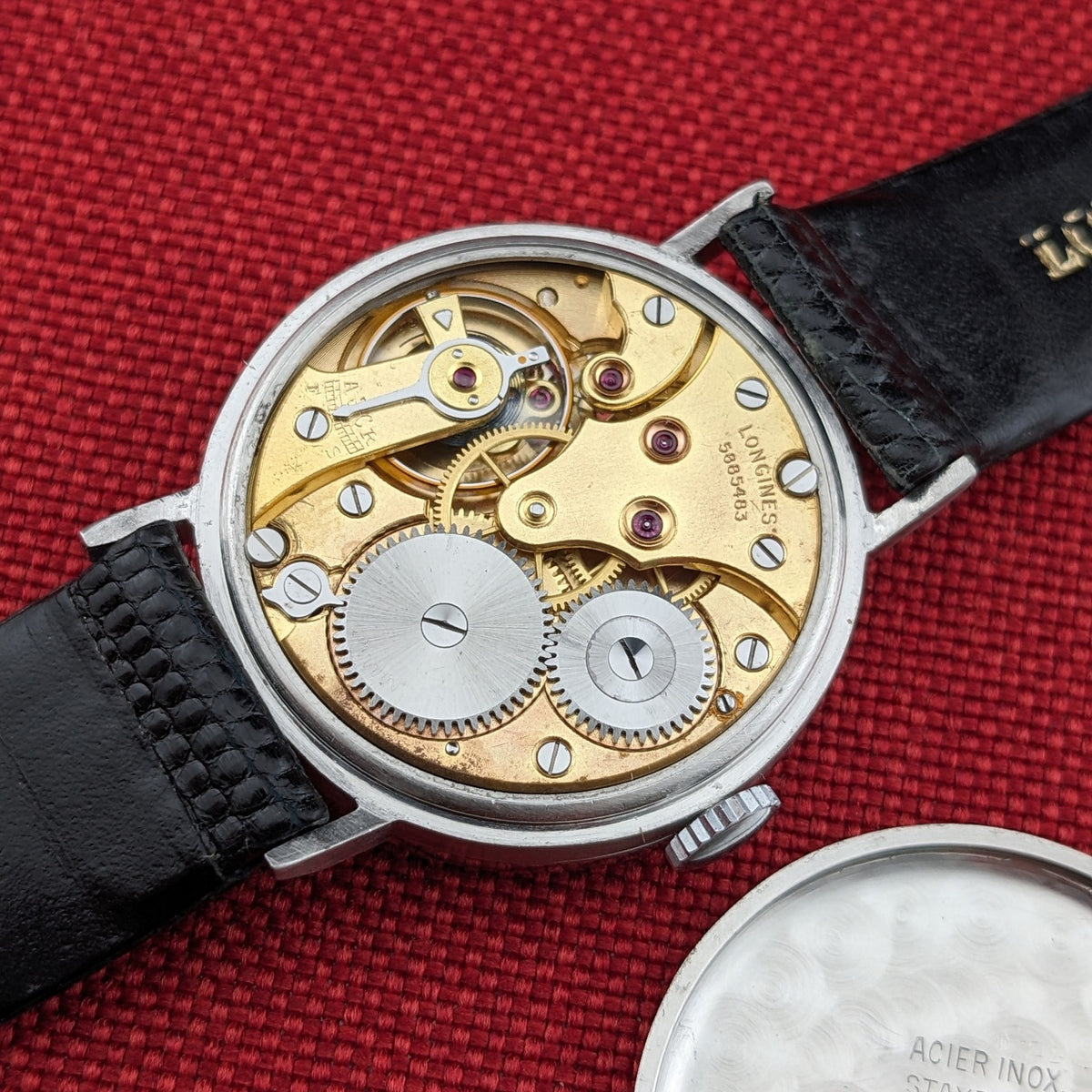 Longines Calatrava reference 20757 shown open on red fabric, with its gold-toned mechanical movement, black strap, and detached round caseback visible.