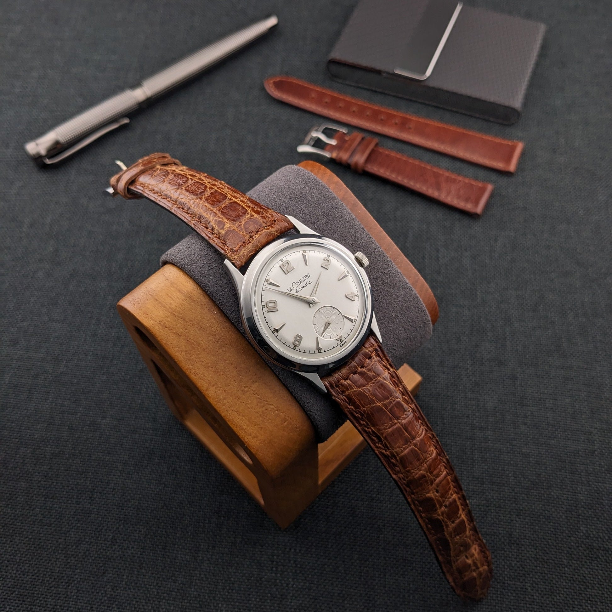 Jaeger-LeCoultre Automatic E328 watch on a wooden stand with a brown strap, spare straps, pen, and black card holder.