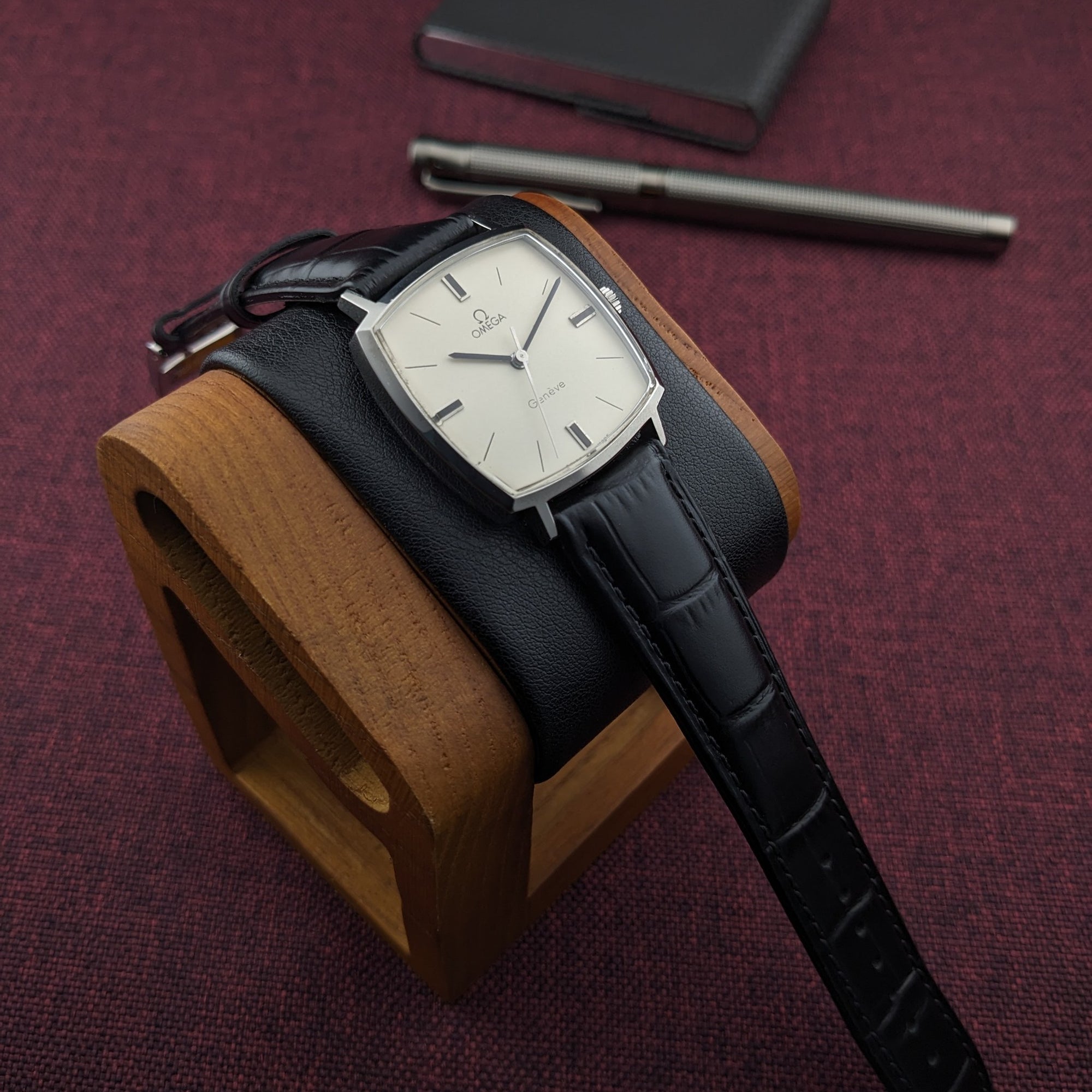 Omega Genève reference 131.022 with a cream square dial and black leather strap, displayed on a wooden watch stand over burgundy fabric.