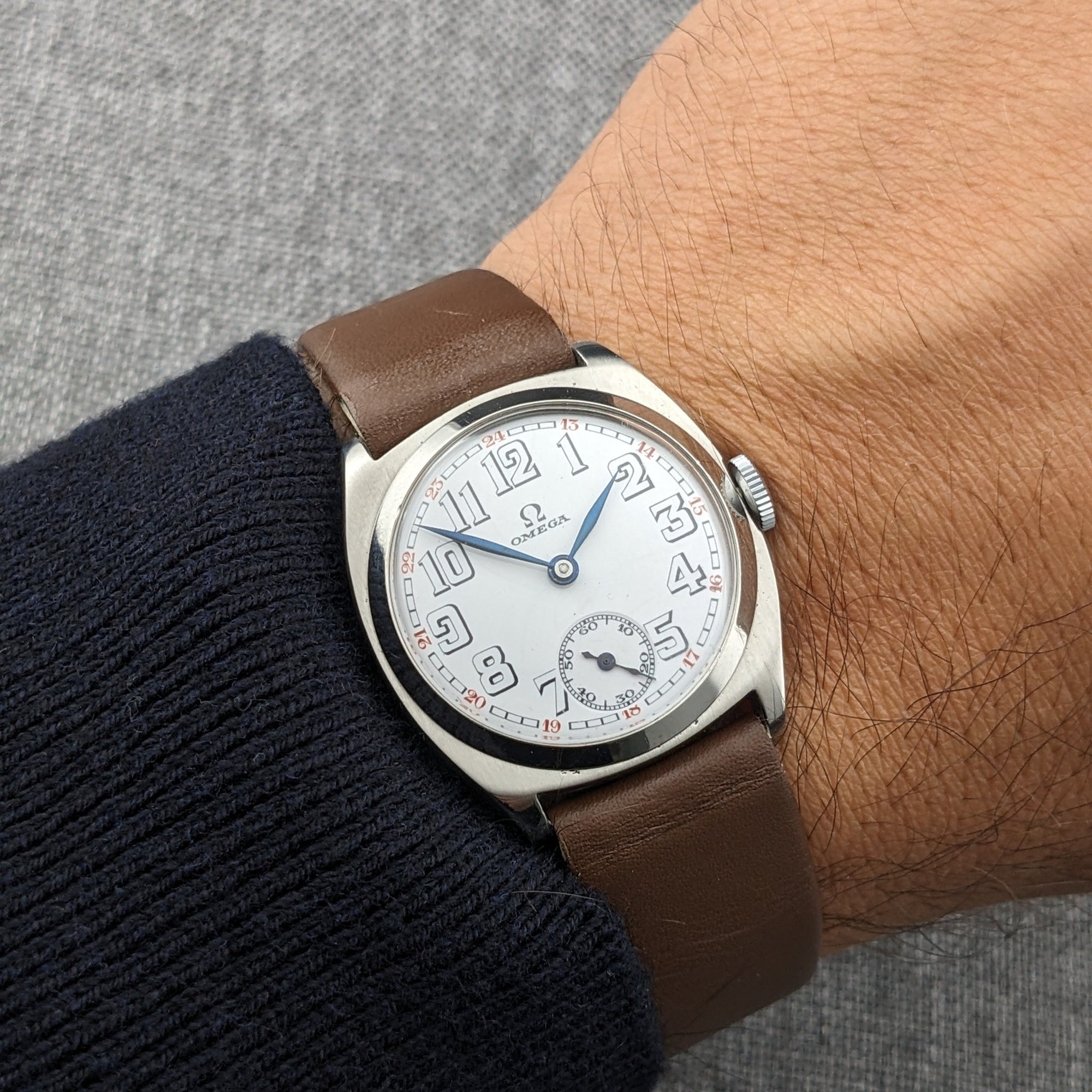 Omega wristwatch with a white dial, oversized Arabic numerals, small seconds subdial, blue hands, and brown leather strap on a wrist.