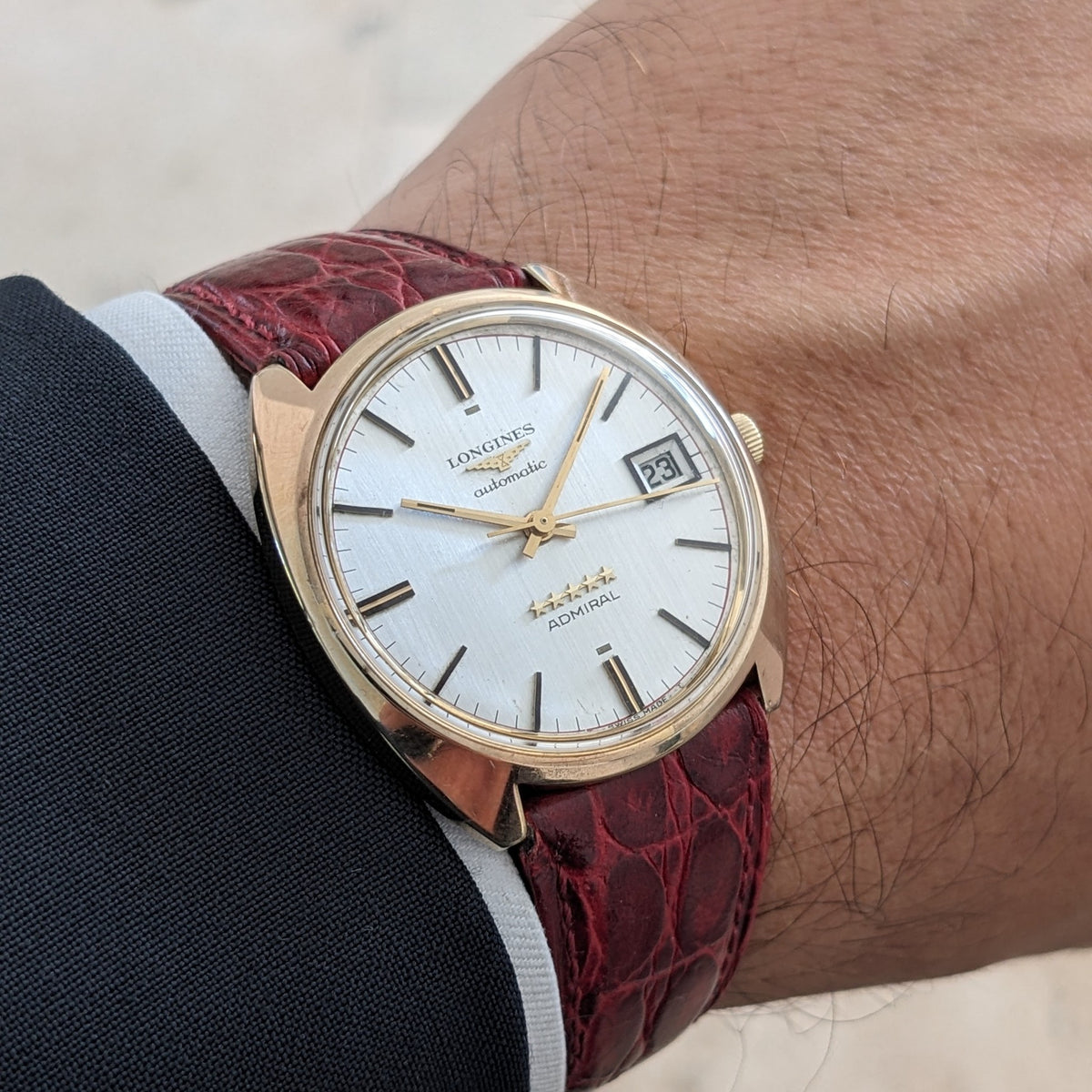 Longines Admiral Referenz 8343-1 am Handgelenk, zeigt ein goldfarbenes rundes Gehäuse, silbernes Zifferblatt, Datumsfenster und burgunderrotes Textilarmband.