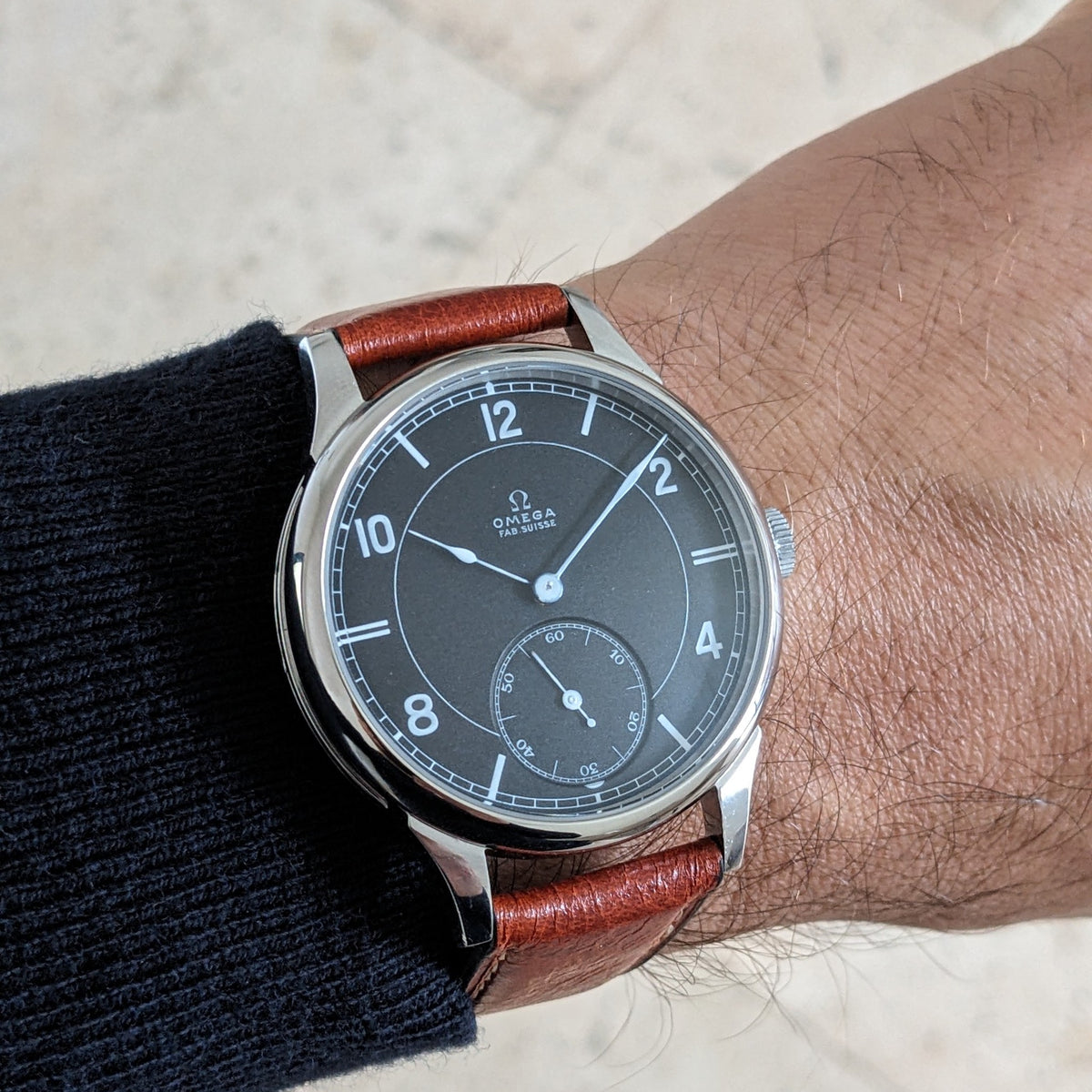 Omega-Armbanduhr mit dunklem Zifferblatt, arabischen Ziffern, kleiner Sekunde, poliertem Gehäuse und braunem Lederarmband am Handgelenk.