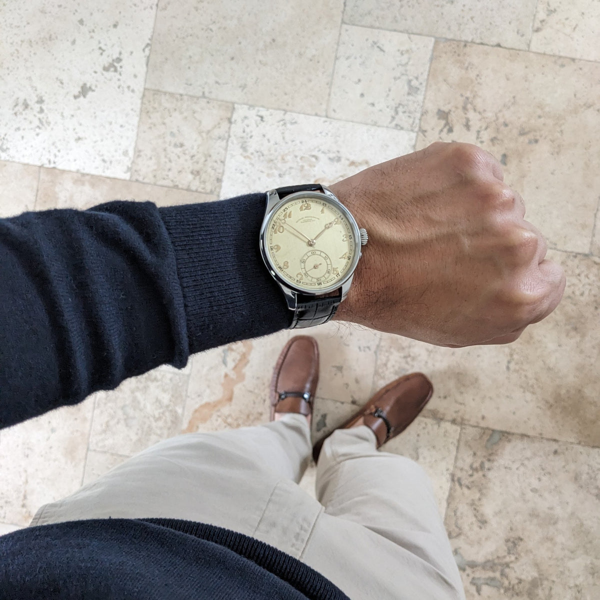 Deutsche Präzisions-Uhrenfabrik Glashütte Armbanduhr mit cremefarbenem Zifferblatt und schwarzem Armband, auf einem Handgelenk über Fliesenboden gezeigt.