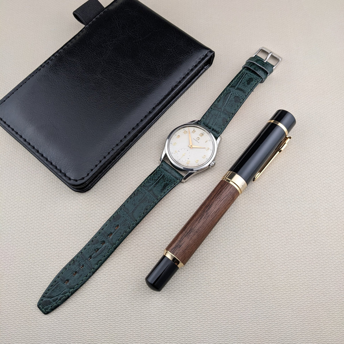 Orologio OMEGA Classic referenza 2639-11 con cinturino verde, custodia in pelle nera e penna in legno e nero disposti diagonalmente su tessuto testurizzato.