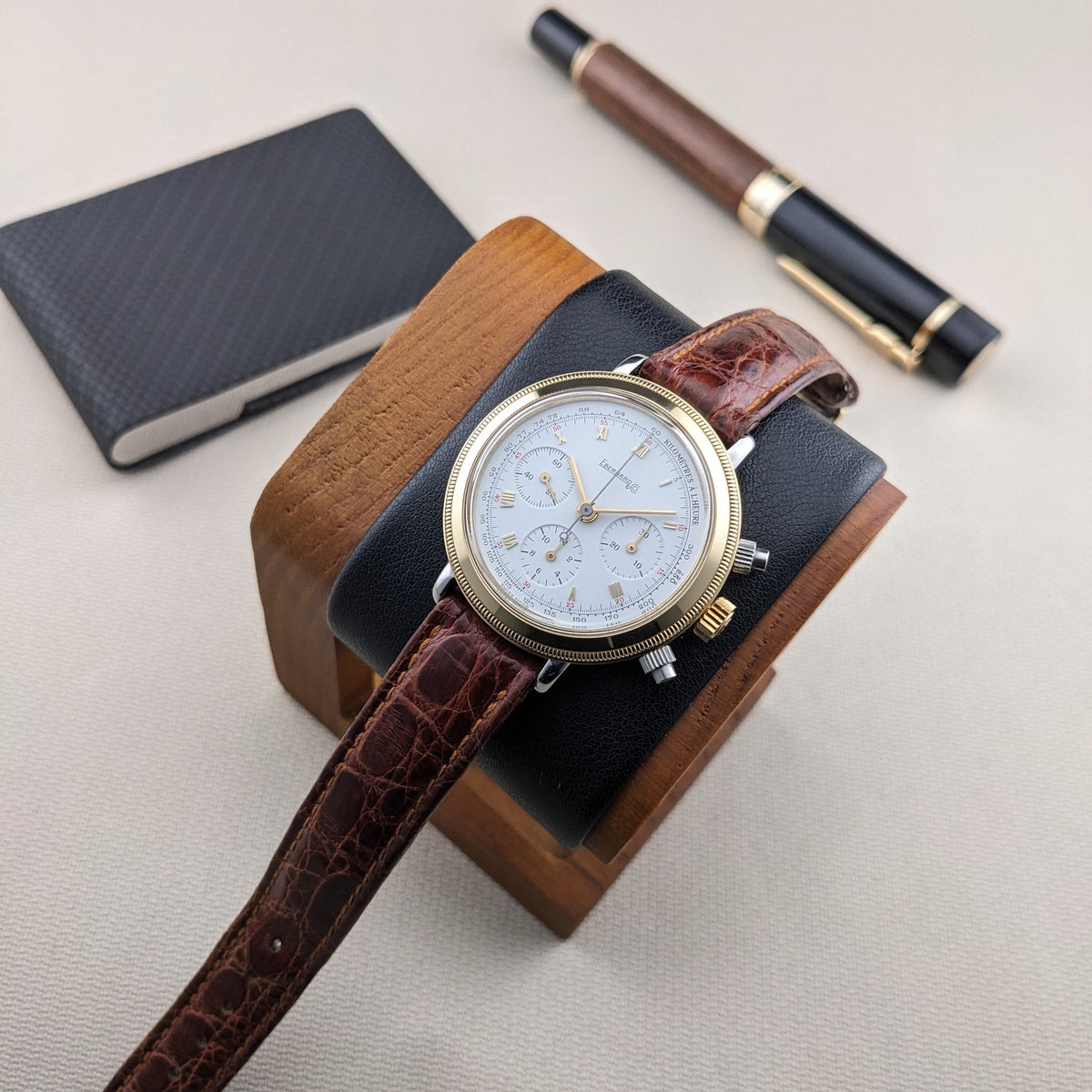 Eberhard & Co.. Godron Chrono reference 32031 rests on a wooden watch stand, with a notebook and fountain pen in the background.