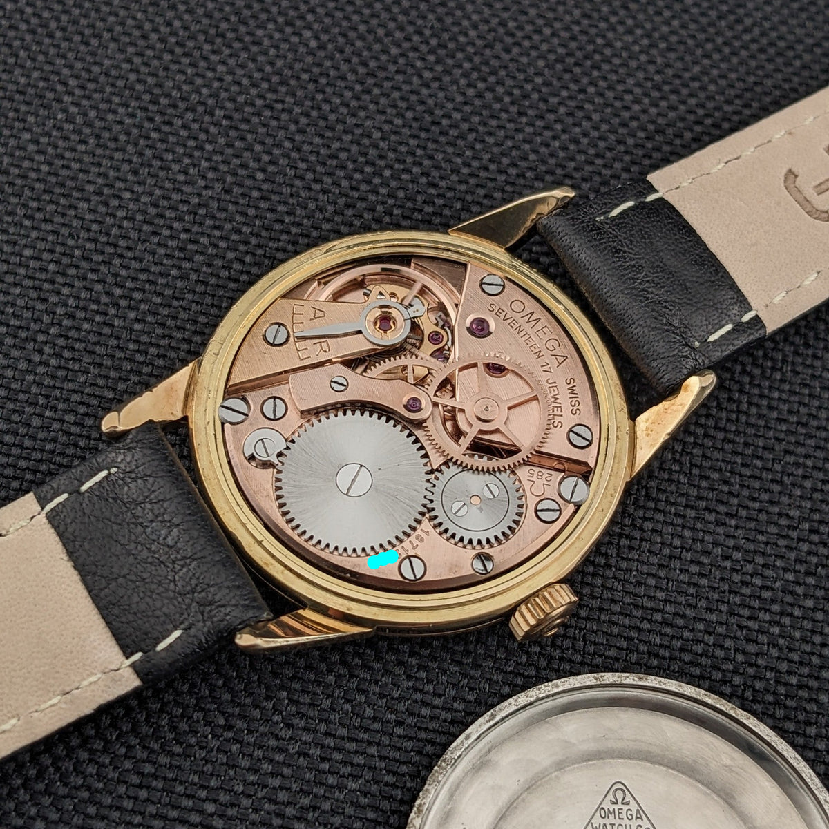 Omega Classic référence 14392-61-SC ouverte, affichant son mouvement mécanique, son bracelet en cuir noir et son fond de boîtier détaché sur tissu texturé.