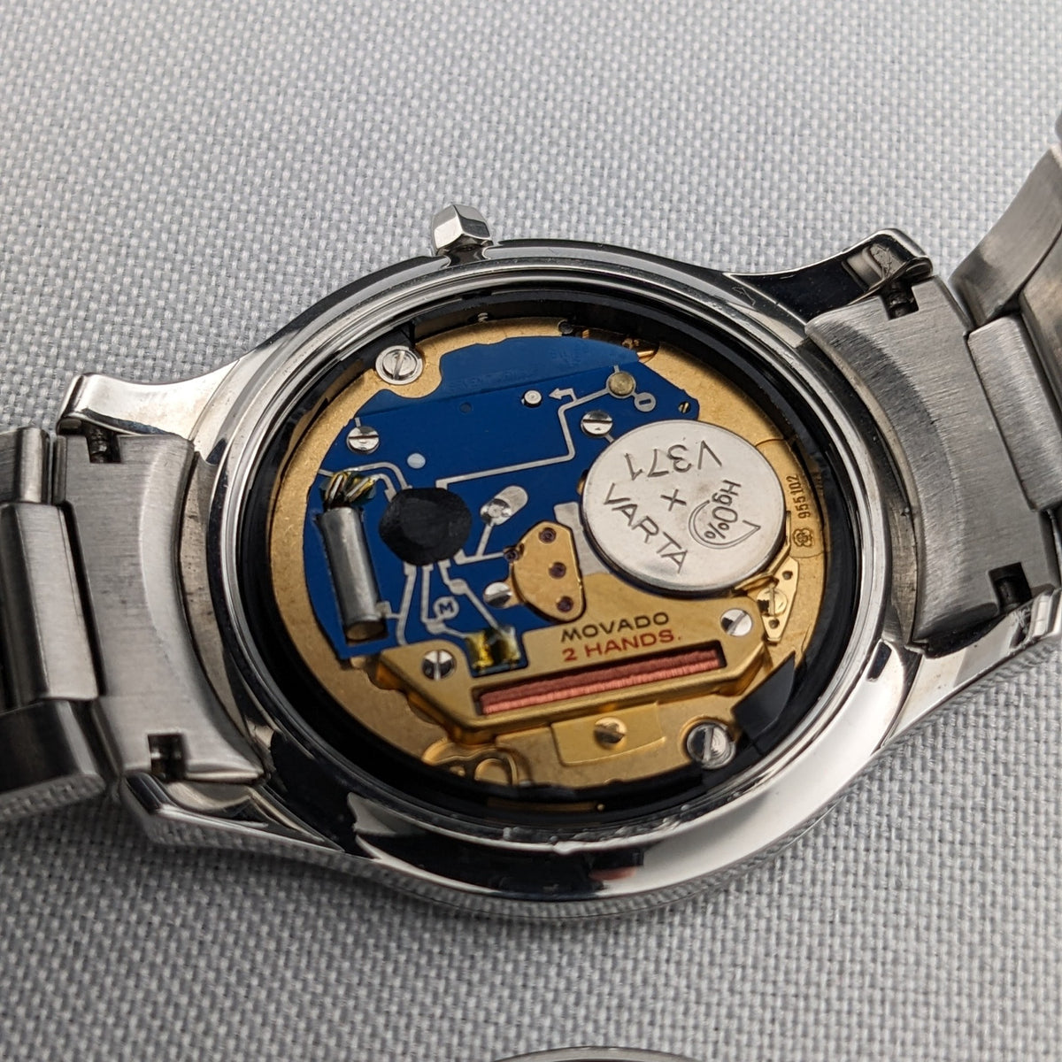 Montre Movado ouverte montrant un mouvement bleu et or, vis visibles, batterie et bracelet entourant le boîtier.
