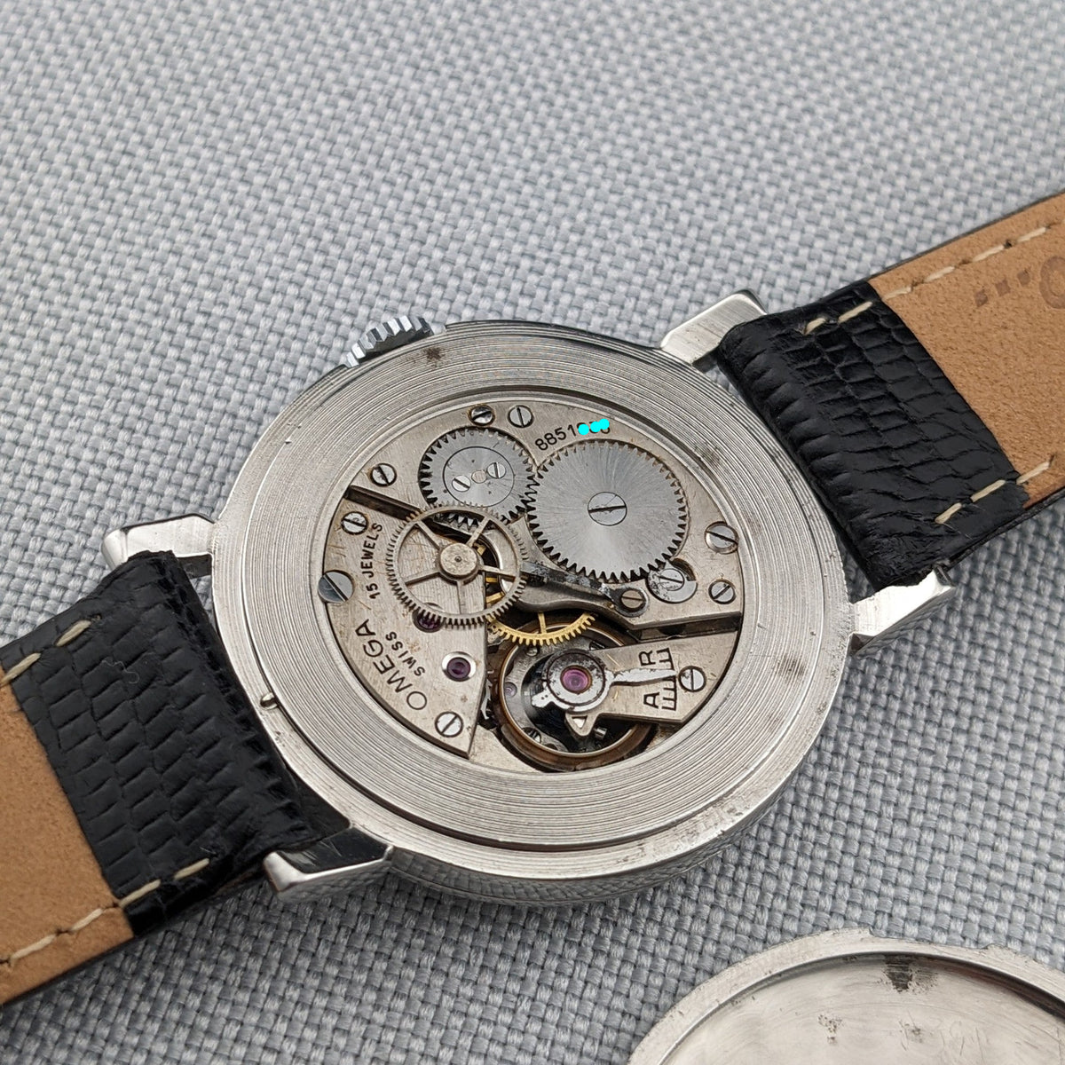 Omega watch opened to reveal its mechanical movement, with gears, bridges and screws visible beside the black strap.
