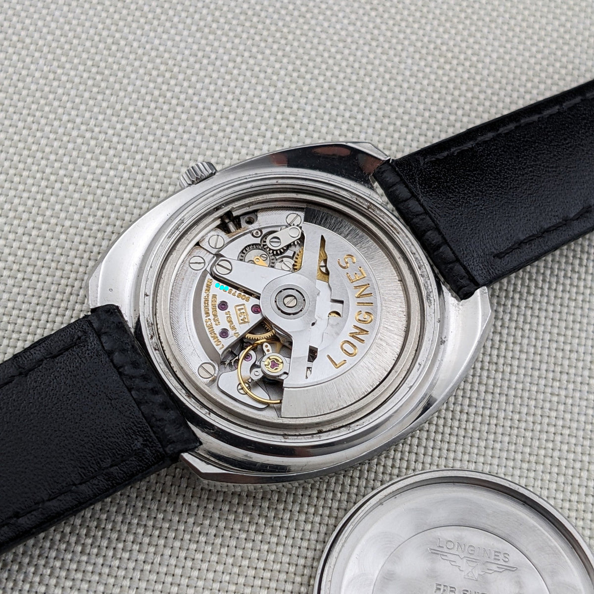 Longines Ultrachron reference 8429 with its automatic movement exposed, black strap attached and detached engraved caseback beside it.