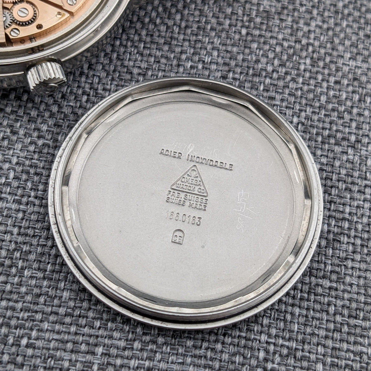 Interior of the Omega Geneve Automatic reference 166.0163 caseback, showing engraved Omega markings and reference text on a gray woven surface.