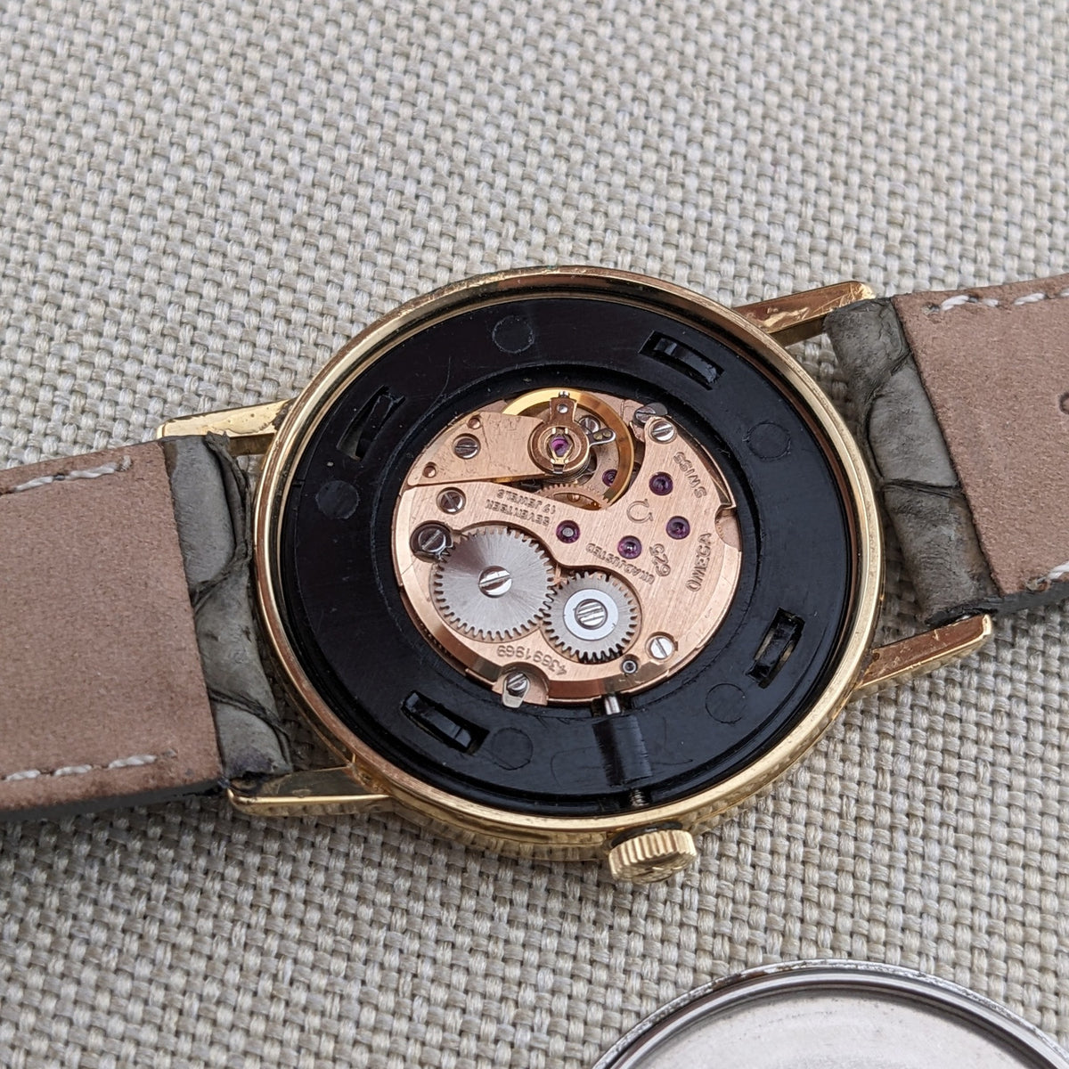 Open Omega De Ville reference 111 0156 showing its mechanical movement, gold-tone case, crown, and gray textured strap.