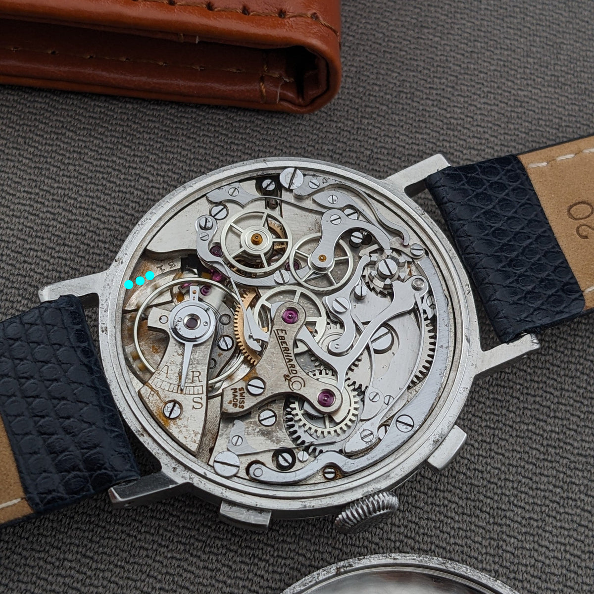 Open Eberhard & Co. Pre-Extra Fort displays its detailed mechanical movement, blue strap, crown and pushers beside a brown wallet.