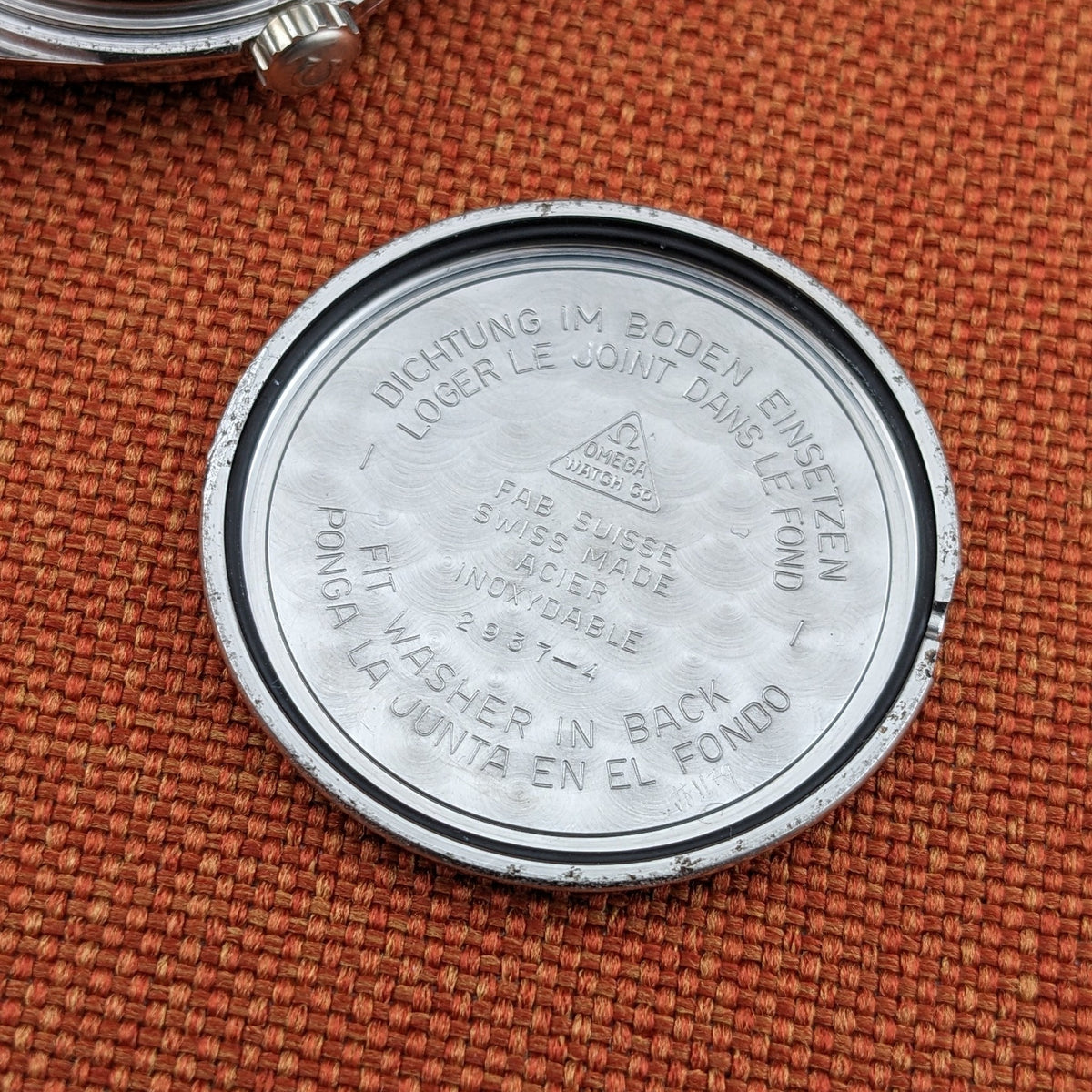 Detached circular caseback for the Omega Seamaster reference 29374, showing engraved text and the Omega emblem against woven orange fabric.
