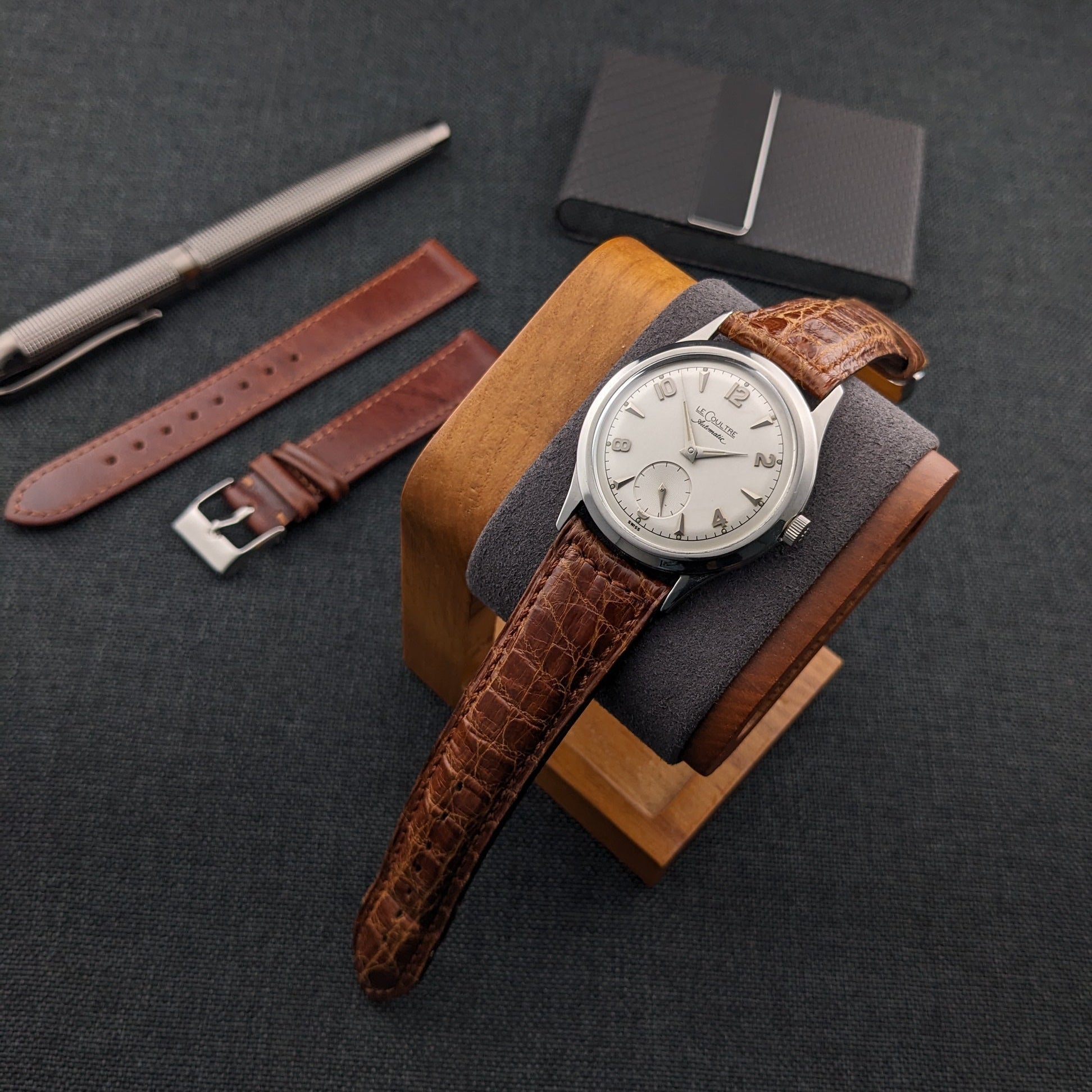 Jaeger-LeCoultre Automatic E328 rests on a wooden display block beside two brown straps, a pen, and a black card holder.