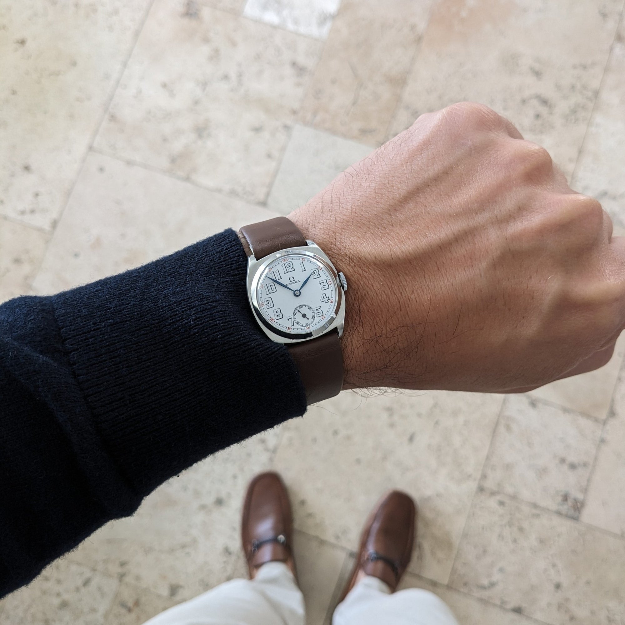 Omega watch shown on a wrist over a tiled floor, with a white numbered dial, brown strap, dark sleeve, trousers, and brown shoes visible.