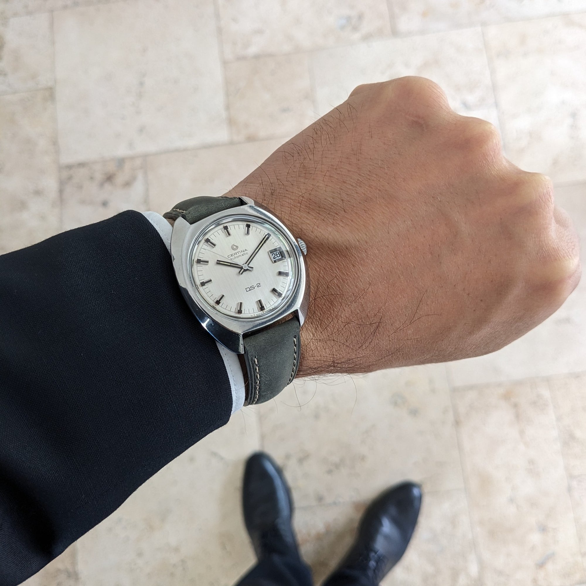Wrist view of the Certina DS-2 Automatic reference 5801 300 with its silver dial and dark leather strap against a tiled floor.