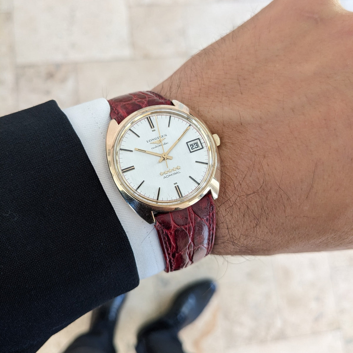 Longines Admiral 8343-1 am Handgelenk neben einem dunklen Anzugärmel, mit sichtbarem silbernen Zifferblatt und burgunderrotem Armband.