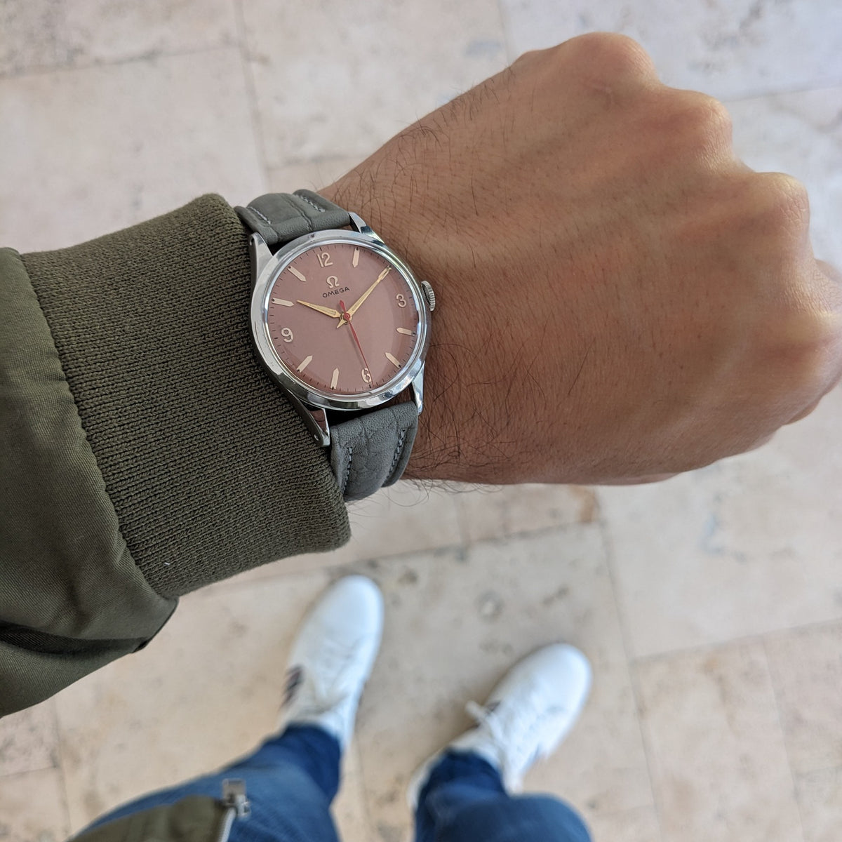 Omega Classic 2760-6 SC auf einem Unterarm von oben, mit rosafarbenem Zifferblatt und grauem Armband auf einem gefliesten Boden.
