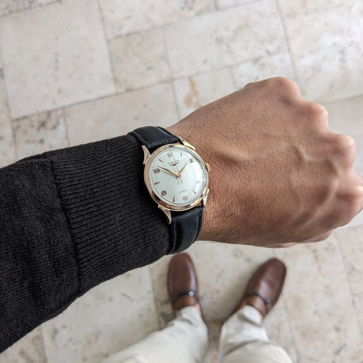 Longines Automatik Referenz 6058 am Handgelenk über Fliesenboden, mit schwarzem Armband und hellem rundem Zifferblatt.