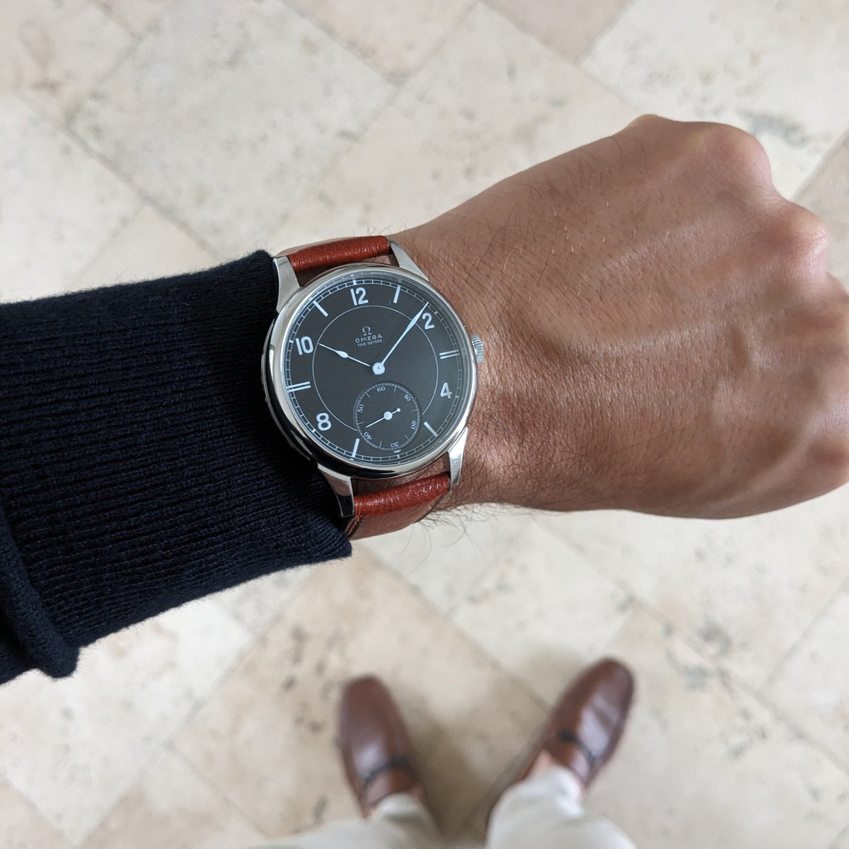 Omega-Armbanduhr von oben am Handgelenk, mit dunklem Zifferblatt und braunem Lederarmband auf hellem Fliesenboden.