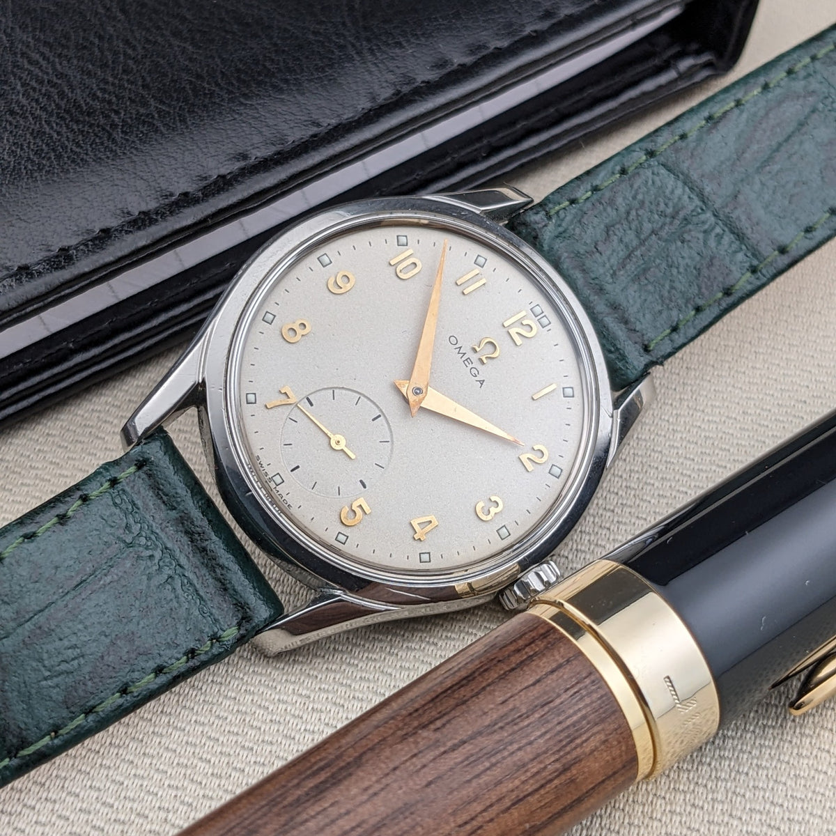 Vista ravvicinata del quadrante argentato OMEGA Classic 2639-11 con numeri arabi dorati, secondi ausiliari, cinturino verde, cassa e penna.