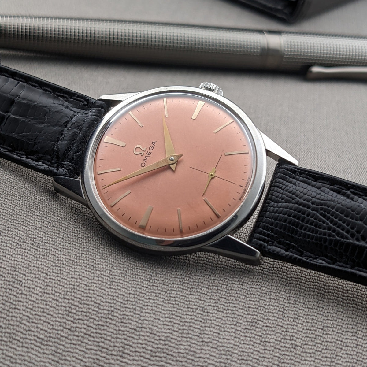 Omega Classic referenza 14391-1 esposto ad angolo con quadrante rosa, cassa argento, cinturino nero e penna metallica su tessuto grigio.