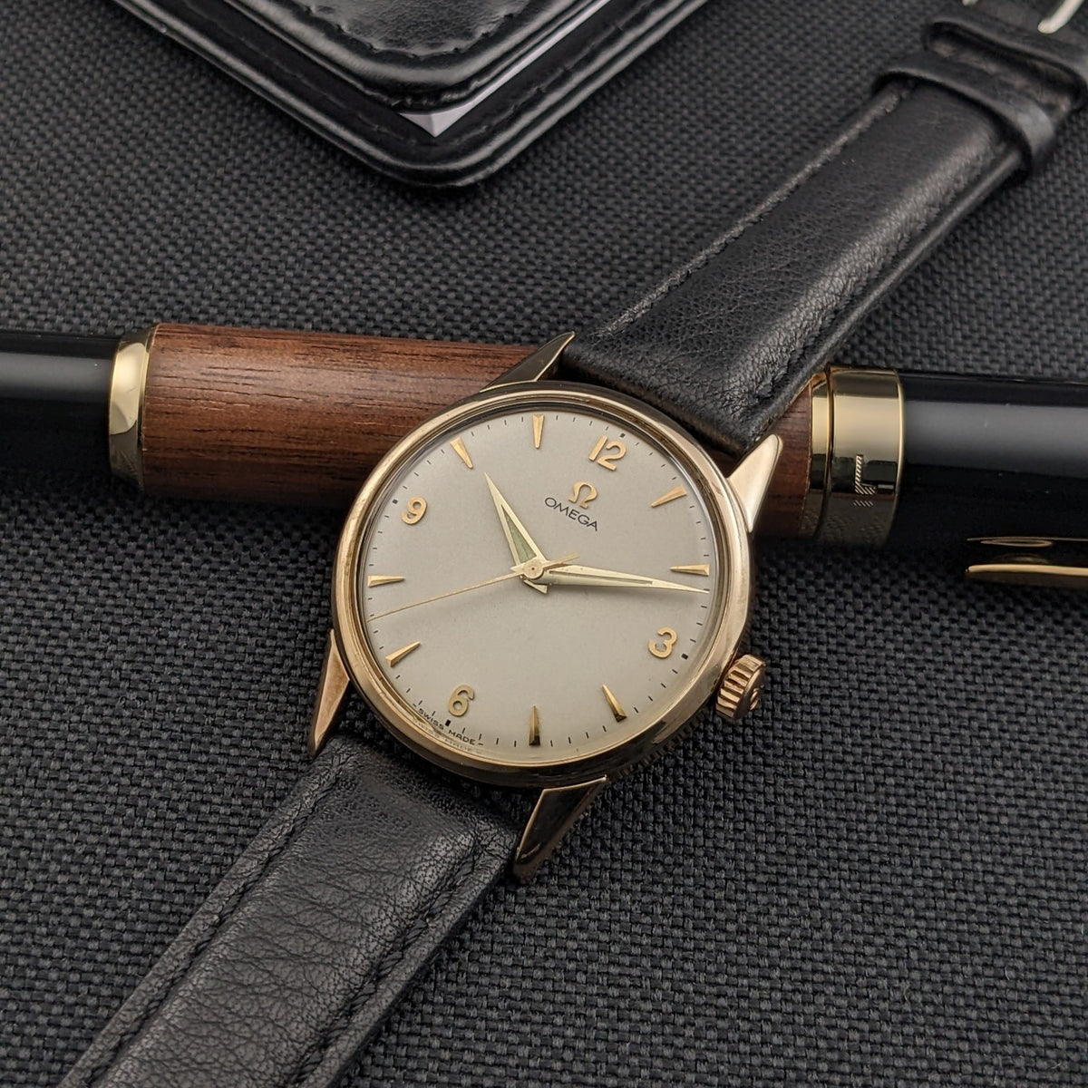 Omega Classic référence 14392-61-SC avec cadran crème, chiffres arabes et index dorés, et bracelet en cuir noir à côté d'un stylo.