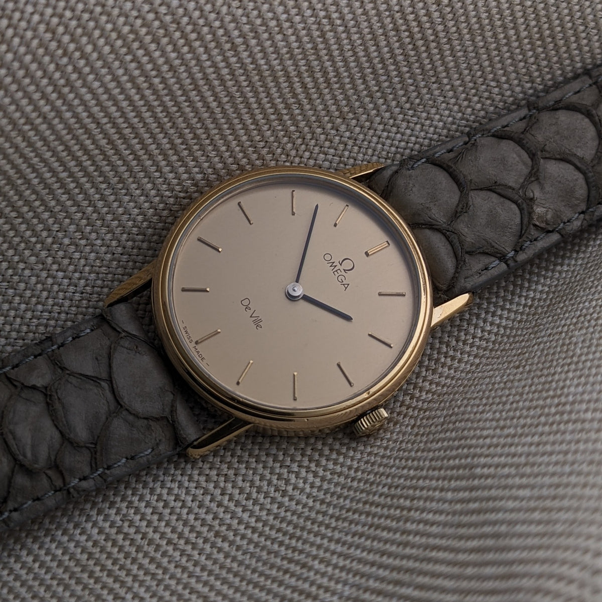Omega De Ville reference 111 0156 with cream dial, slim baton markers, blue hands, gold-tone case, and gray crocodile-pattern strap.