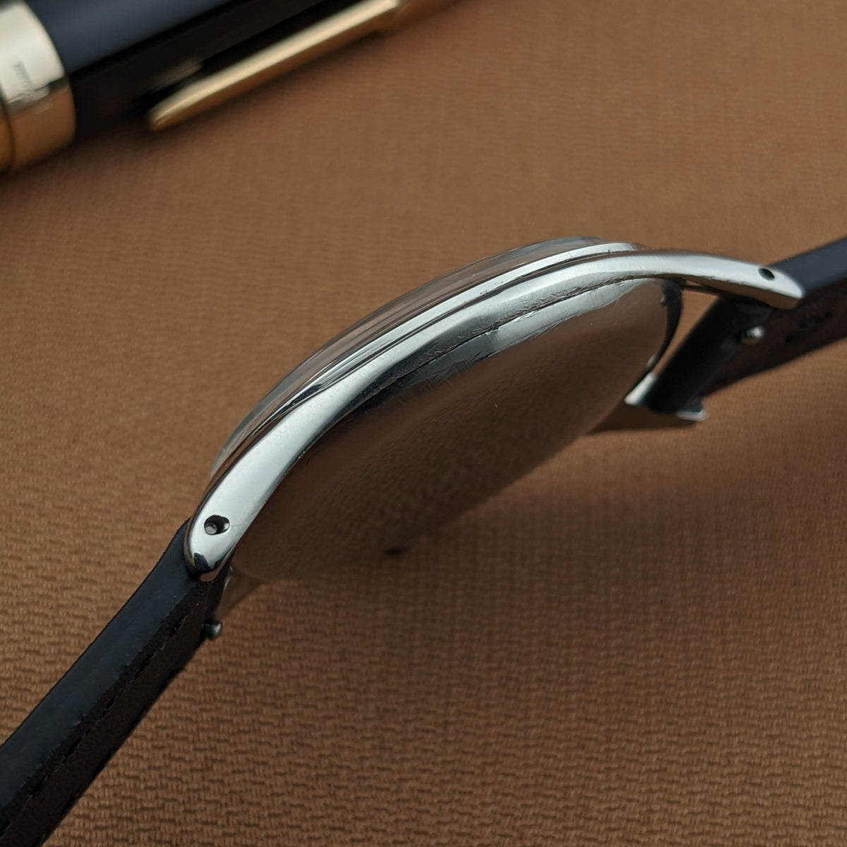 Side view of the Omega Jumbo reference 2505-5, showing its curved polished case, lugs, black leather strap, and wooden pen.