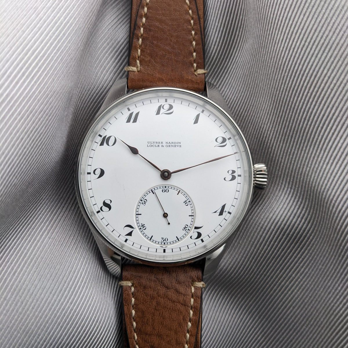 Ulysse Nardin Locle & Genève कलाई घड़ी भूरे चमड़े के पट्टे पर खड़ी ग्रे कपड़े के खिलाफ खड़ी।