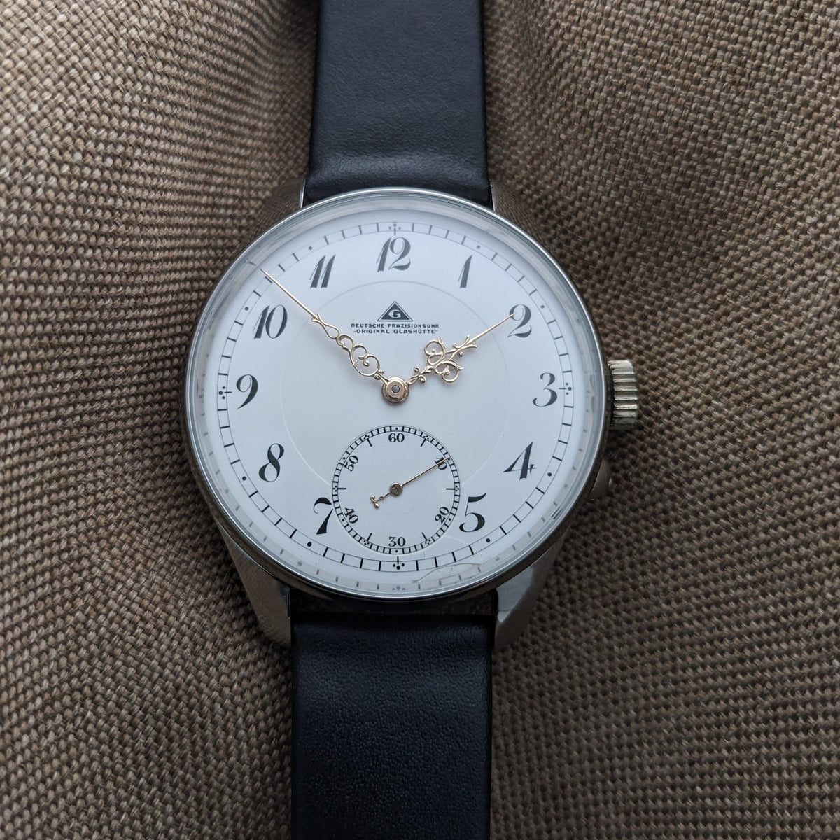 Vollansicht der Deutsche Präzisionsuhr „Original Glashütte” auf einem schwarzen Lederarmband, arrangiert gegen textur