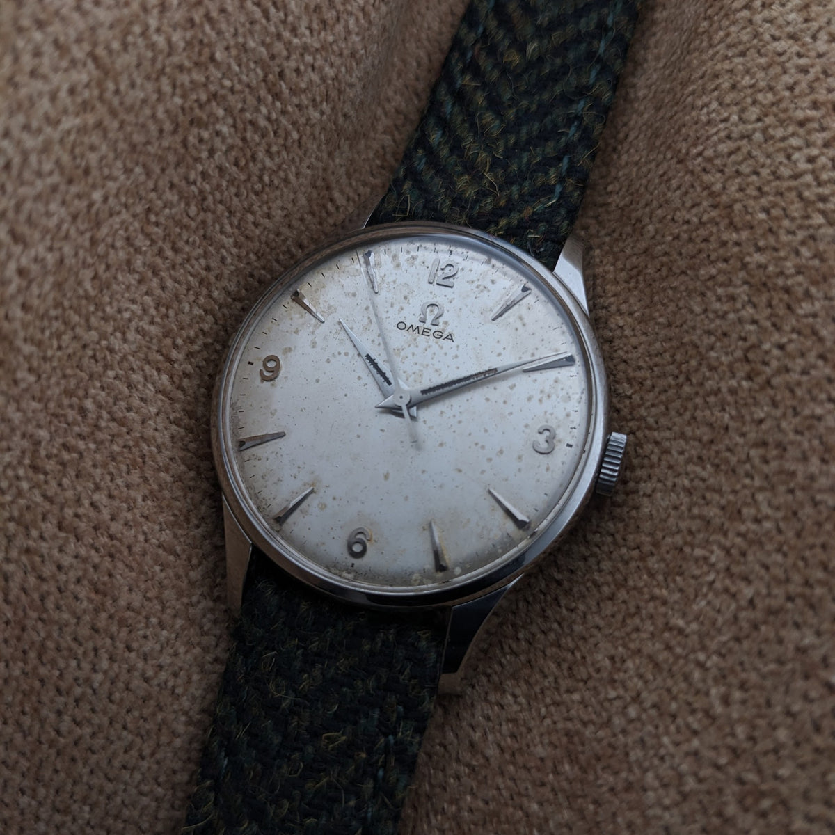 Omega Classic Referenz 2810-2 SC mit hellem Zifferblatt, arabischen Ziffern, aufgesetzten Stundenmarkierungen und grünem gemustertem Armband auf gewebtem Stoff.