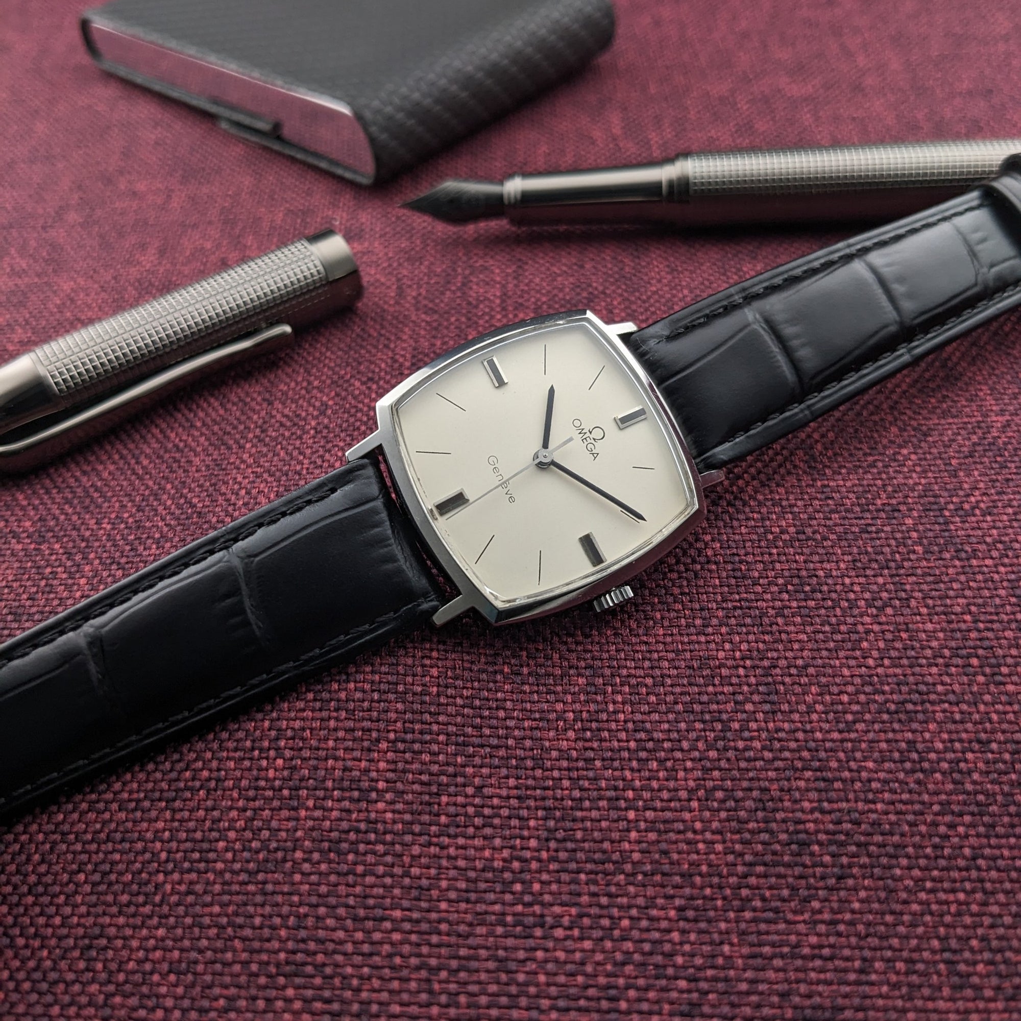 Omega Genève reference 131.022 laid diagonally on burgundy fabric, with a cream dial, black strap, and two silver writing instruments nearby.
