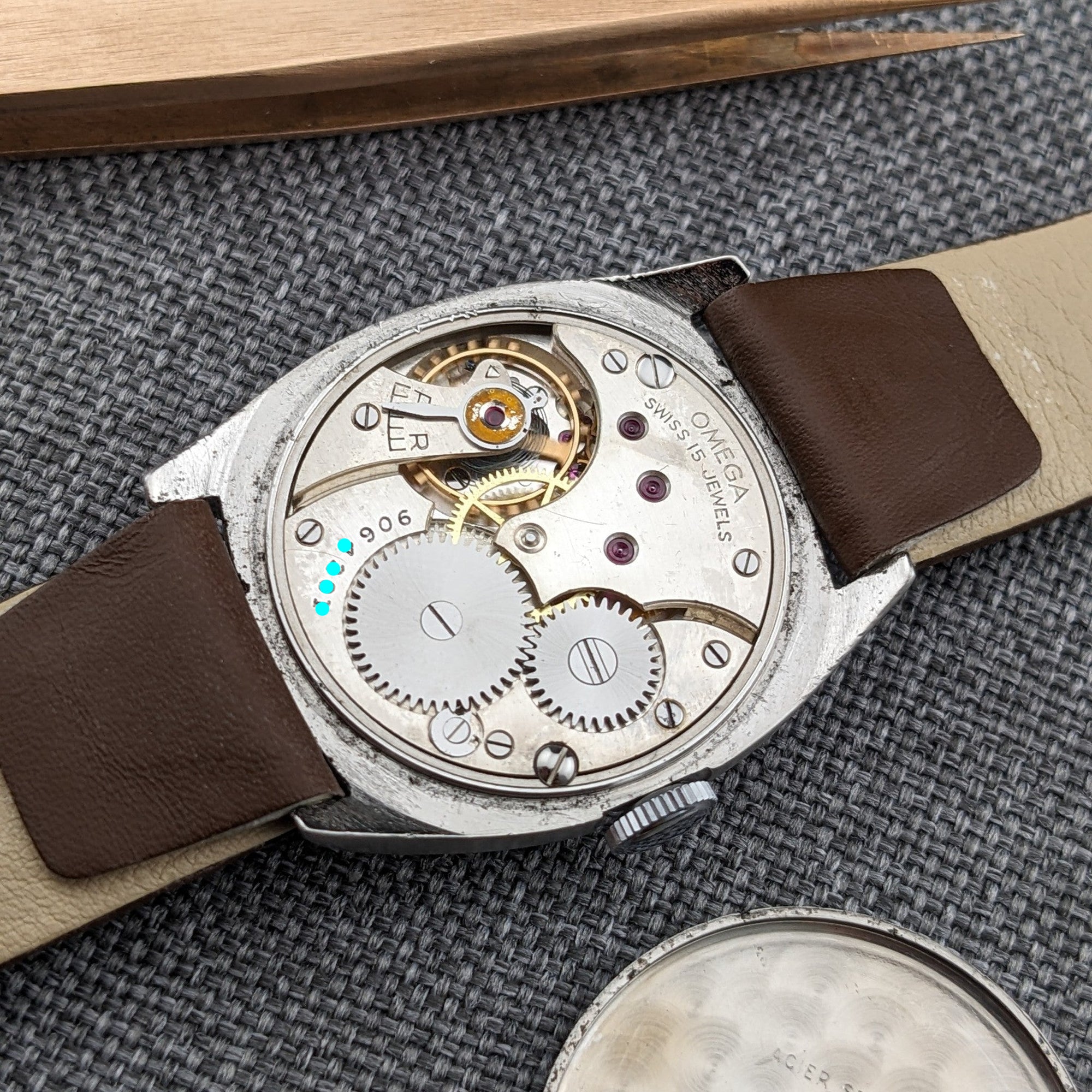 Omega watch viewed from the back with its mechanical movement, gears, jewels, brown strap, and detached round caseback arranged on gray fabric.