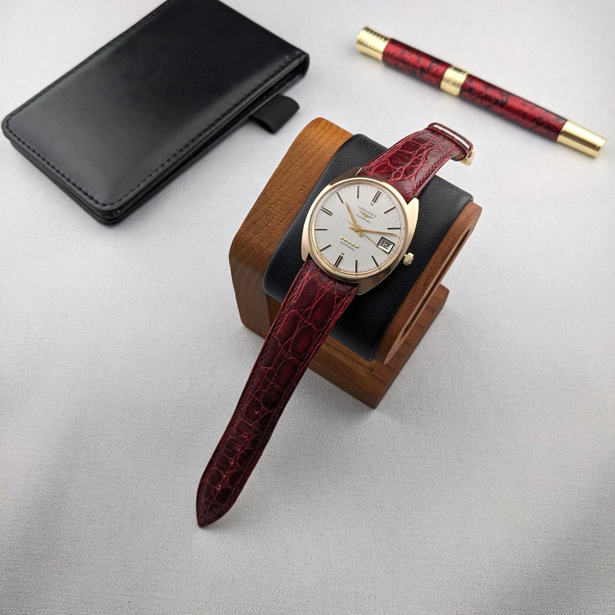 Longines Admiral Referenz 8343-1 auf einem Holzuhrenständer neben einem schwarzen Etui und einem burgunder-goldenen Stift.